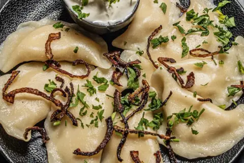 Pyszne pierogi z ziemniakami i boczkiem, posypane chrupiącą cebulką i natką pietruszki. Idealne z kwaśną śmietaną.